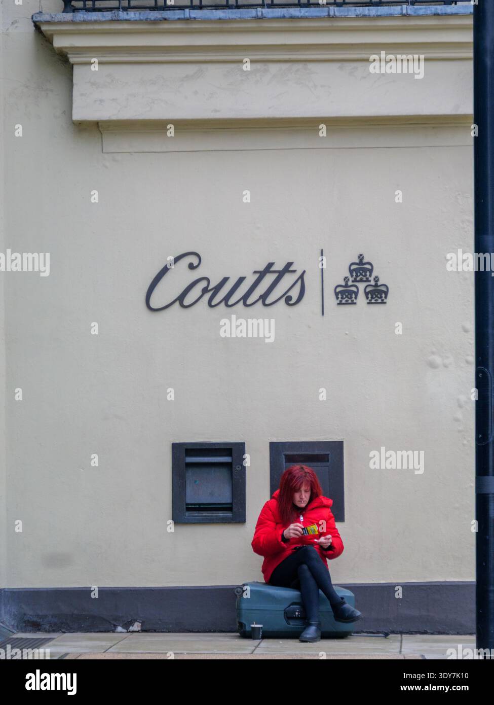 Una donna siede sulla sua valigia per fare una sigaretta arrotolata direttamente sotto il prestigioso emblema delle tre corone di Coutts & Co. Al 440 The Strand, Londra, nel febbraio 2026. Questa sorprendente giustapposizione cattura il forte divario socio-economico della capitale britannica: Un viaggiatore che riposa sul marciapiede sullo sfondo di una delle banche private più esclusive al mondo, che notoriamente richiede ai clienti di avere almeno un milione di sterline in beni investibili. Foto Stock
