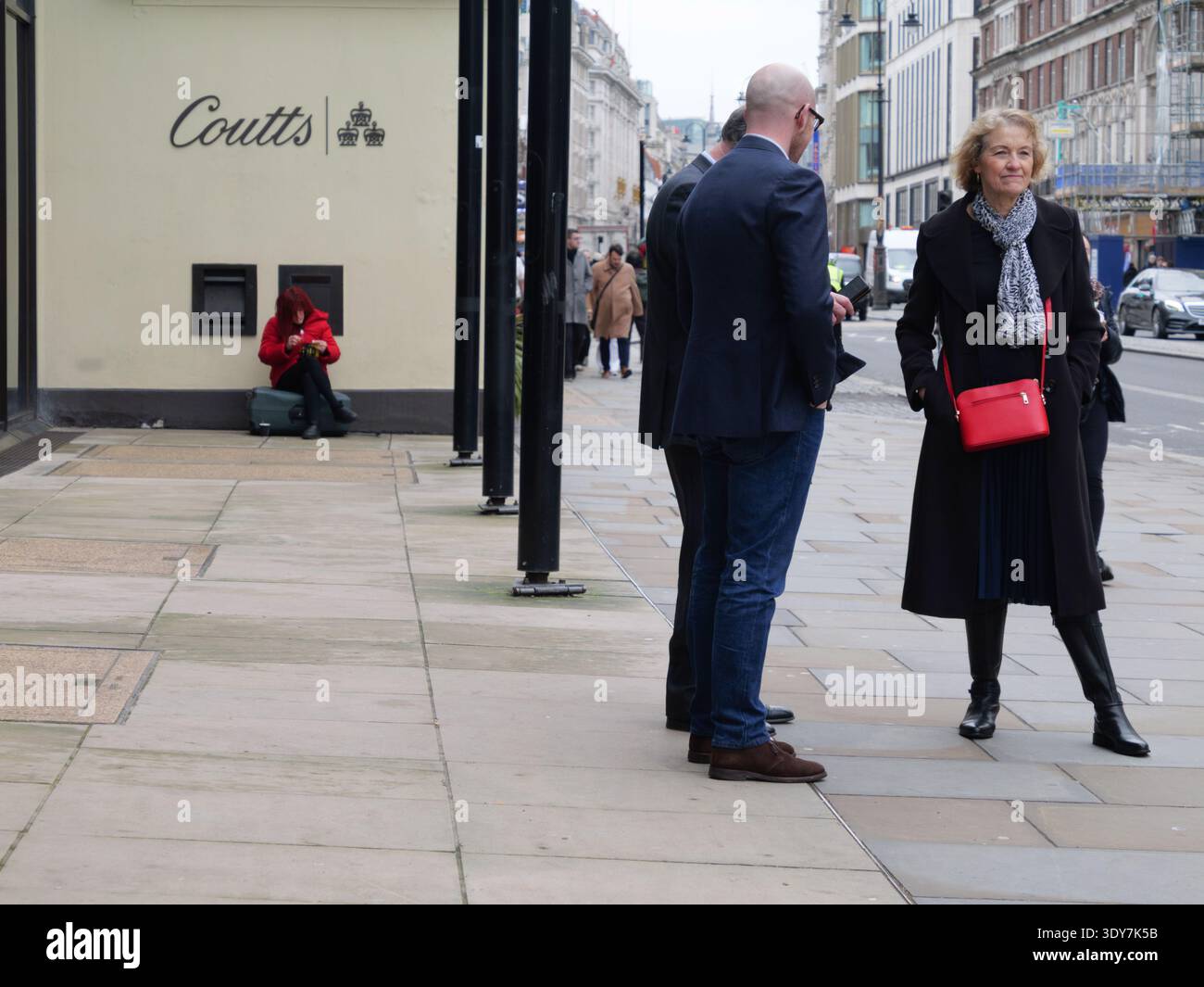 Professionisti ben vestiti e adatti si riuniscono fuori dalla prestigiosa sede di Coutts & Co. Al 440 di The Strand, Londra, mentre una donna siede sulla sua valigia a fare una sigaretta arrotolata direttamente sotto lo storico emblema delle tre corone della banca. Catturata nel febbraio 2026, questa scena presenta un'impressionante giustapposizione del divario socio-economico nella capitale britannica. Il mondo del private banking, che richiede ai clienti di detenere almeno 1 milione di sterline in asset investibili, incontra la realtà della vita di strada transitoria in una delle strade più trafficate di Londra Foto Stock