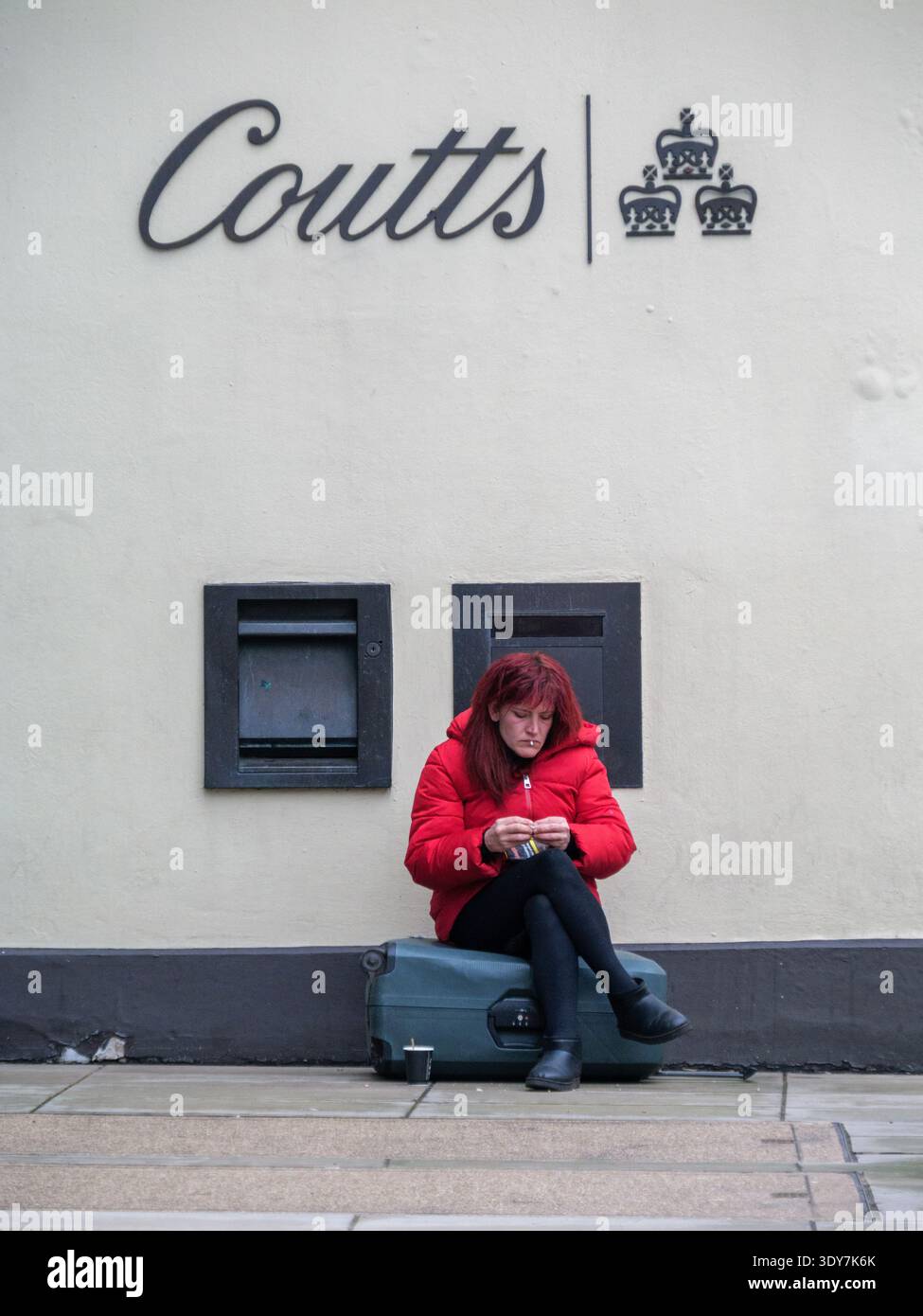 Una donna siede sulla sua valigia per fare una sigaretta arrotolata direttamente sotto il prestigioso emblema delle tre corone di Coutts & Co. Al 440 The Strand, Londra, nel febbraio 2026. Questa sorprendente giustapposizione cattura il forte divario socio-economico della capitale britannica: Un viaggiatore che riposa sul marciapiede sullo sfondo di una delle banche private più esclusive al mondo, che notoriamente richiede ai clienti di avere almeno un milione di sterline in beni investibili. Foto Stock