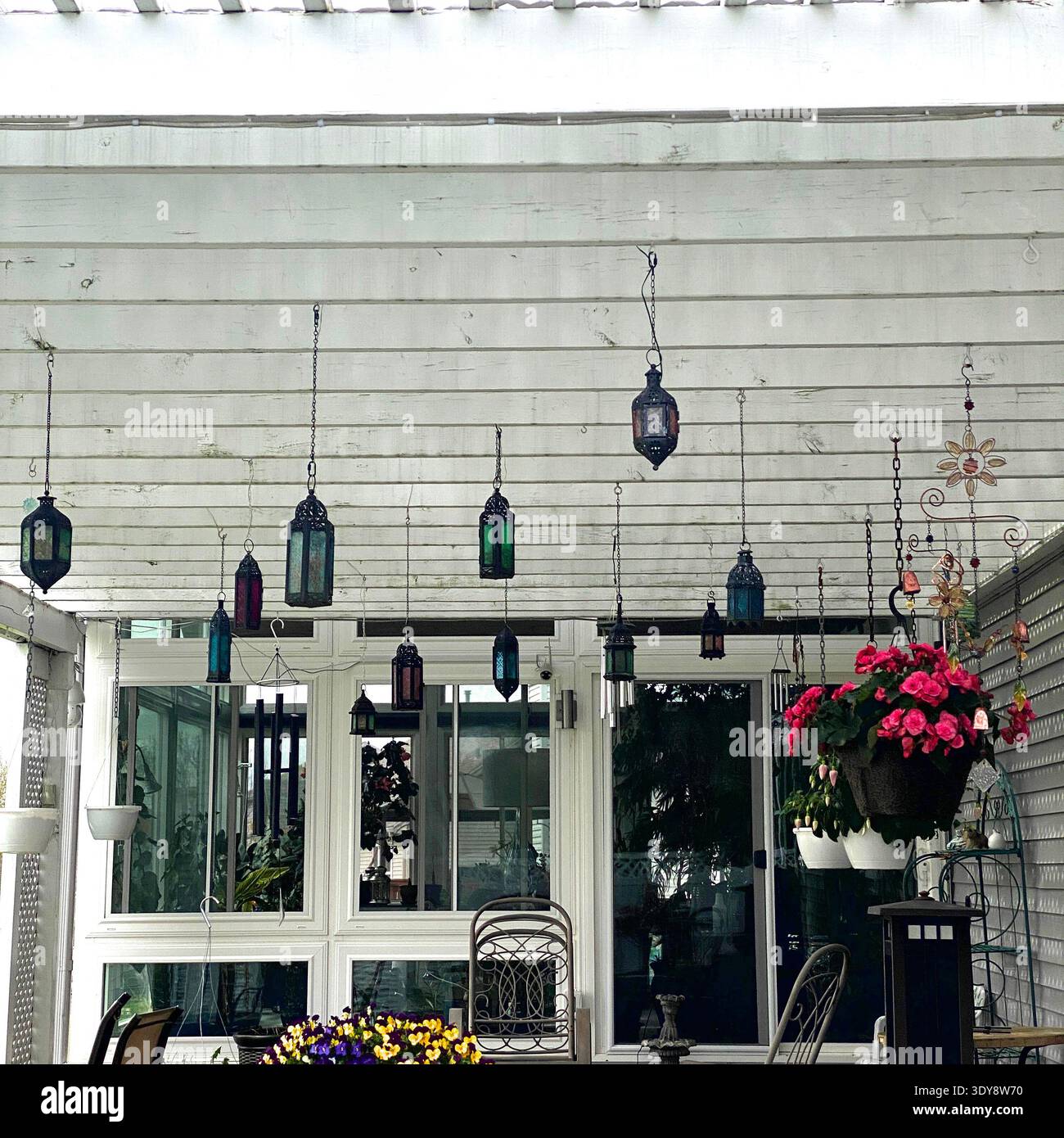 Numerose lampade marocchine appese da un pergolato di legno bianco, adiacente ad un solarium, lungo il retro di una casa, con fiori e campanelli a vento - Immagine stock catturata con smartphone