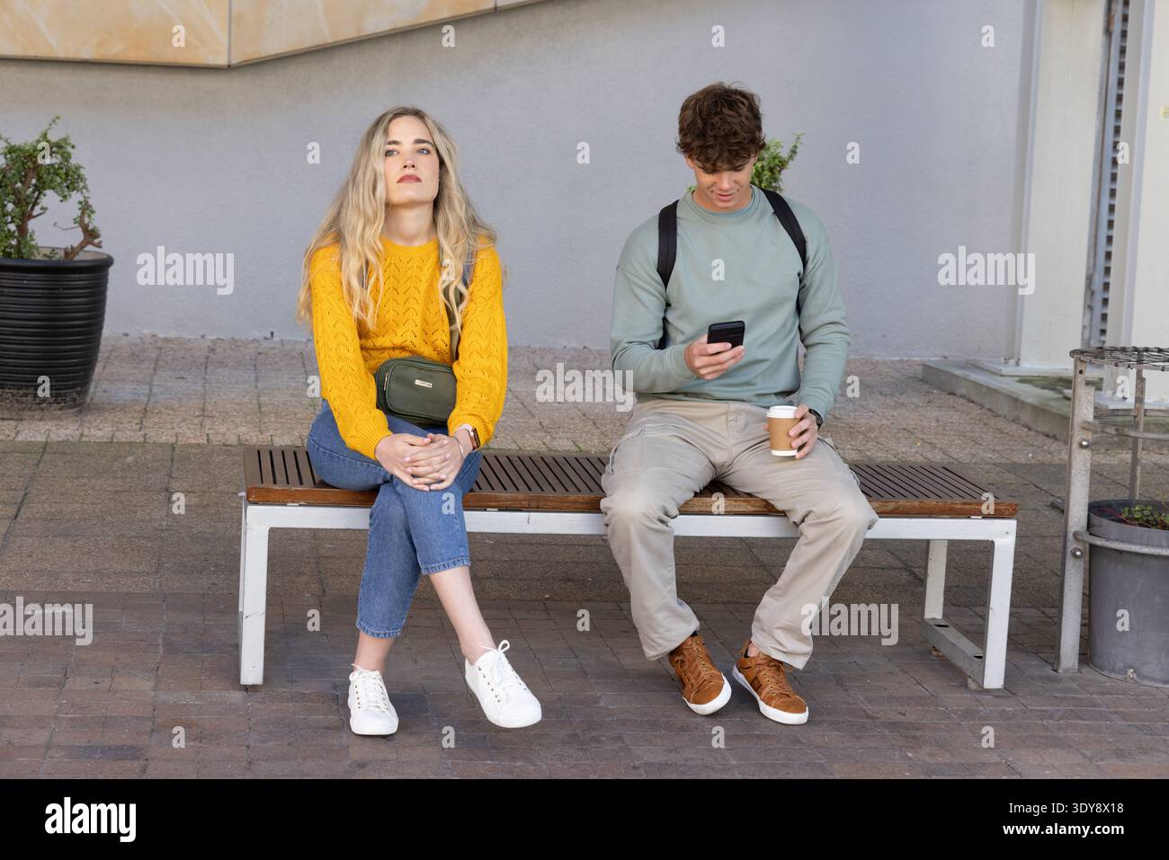 Due persone sedute su una panchina di legno al plaza indossano senape e verde tenendo smartphone e caffè Foto Stock
