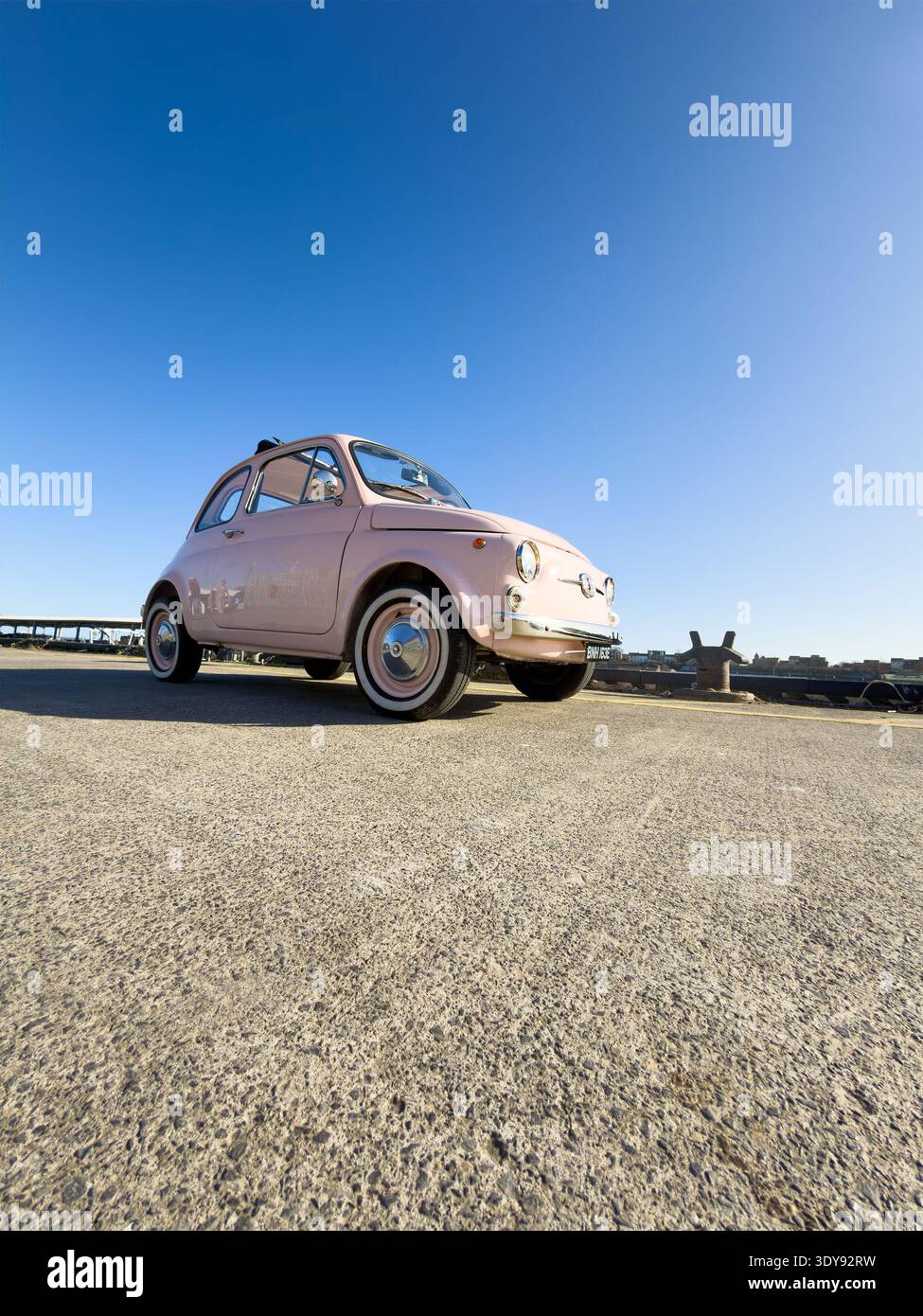 Vista grandangolare bassa di una classica Fiat 500 vintage degli anni '60 rosa pallido parcheggiata su pavimentazione in asfalto vicino al bordo del porto a North Shields Fish Quay, UK - Immagine stock catturata con smartphone