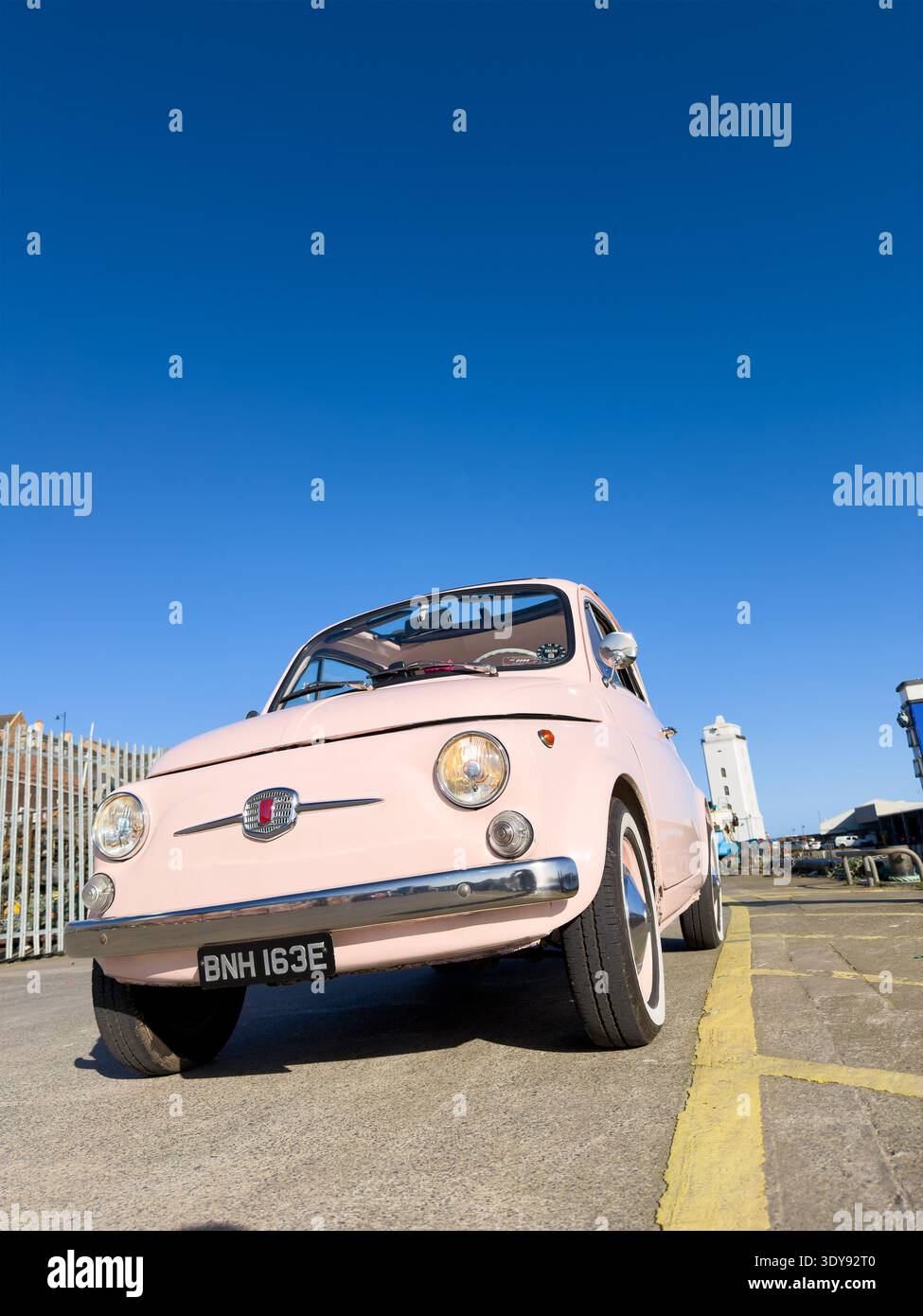 Vista grandangolare bassa di una classica Fiat 500 vintage degli anni '60 rosa pallido parcheggiata su pavimentazione in asfalto vicino al bordo del porto a North Shields Fish Quay, UK - Immagine stock catturata con smartphone