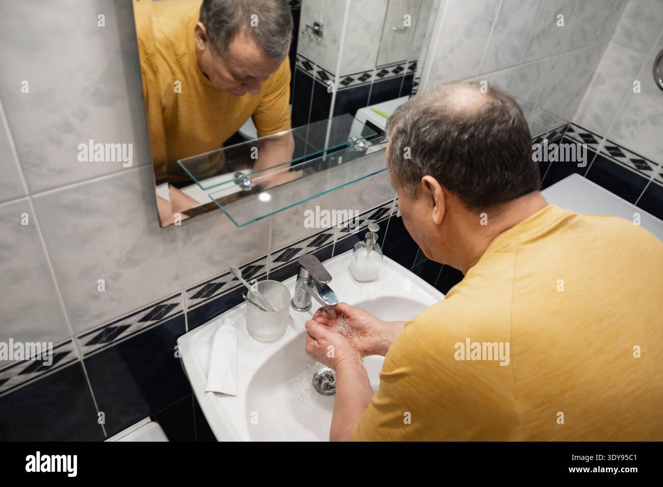 Uomo maturo che lava le mani sotto l'acqua corrente nel lavandino del bagno durante la routine quotidiana di igiene a casa. Routine mattutina. Foto Stock
