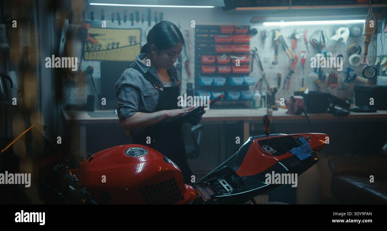La donna asiatica in officina utilizza il tablet per seguire le istruzioni tecniche per il collegamento dei cavi della batteria della motocicletta. Riparazione professionale di motociclette fai da te. Concetto di tecnologia e competenze meccaniche. Foto Stock
