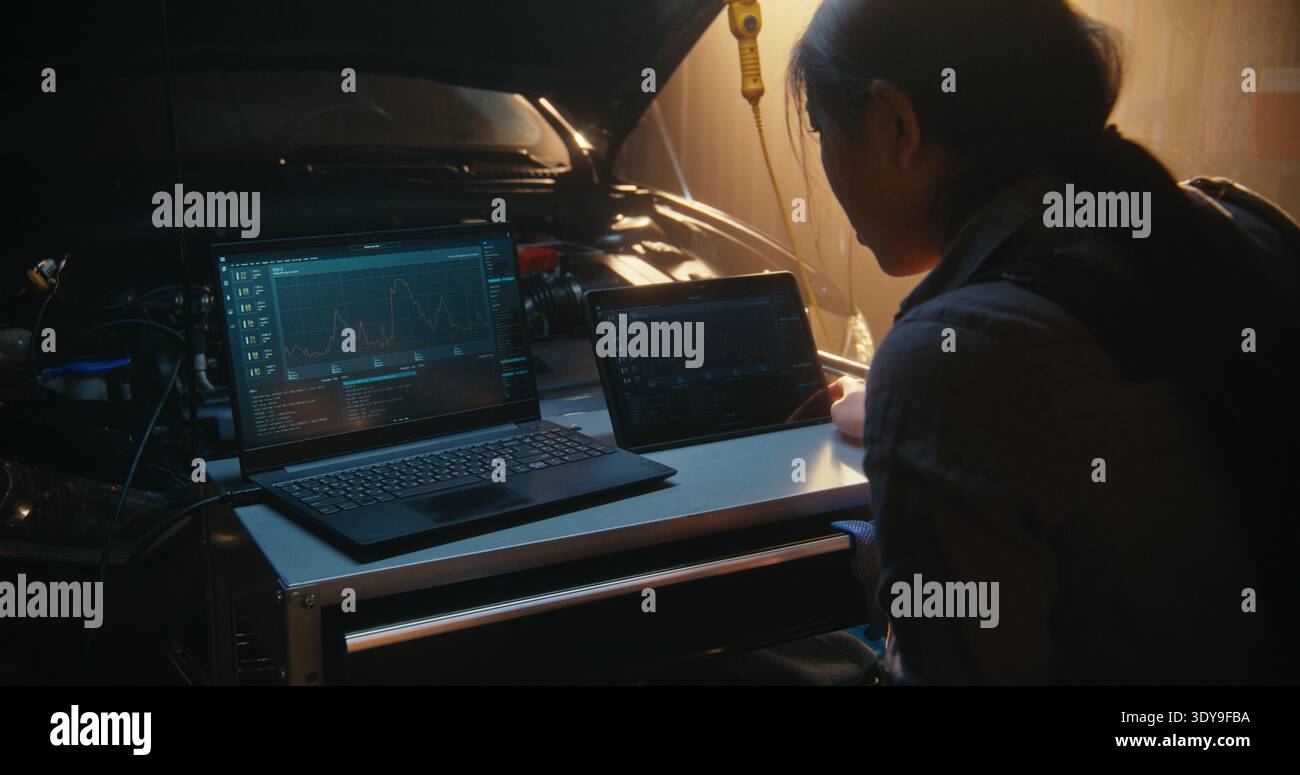 Software di diagnostica sincronizzata da un esperto automatico femminile su notebook e tablet. La giovane donna asiatica gestisce la workstation digitale, monitorando le metriche di salute dei veicoli tramite applicazioni di diagnostica avanzata. Foto Stock