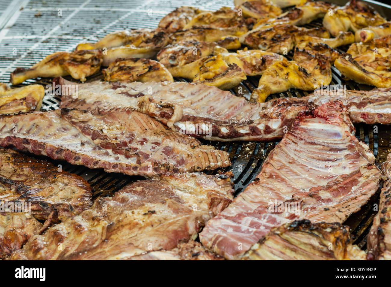 Una grande varietà di costolette di maiale condite e pezzi di pollo arrostiti su una griglia in metallo professionale all'aperto in un mercato alimentare. Foto Stock