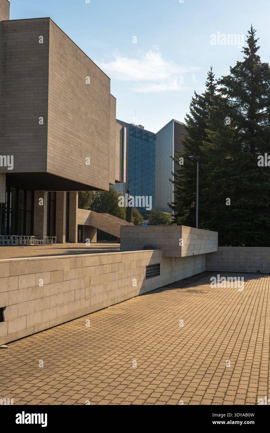 Edificio modernista in cemento illuminato dal sole con volumi a sbalzo e terrazza in pietra che sorge su alti sempreverdi, una statua solitaria sull'asse, con alti edifici in vetro Foto Stock