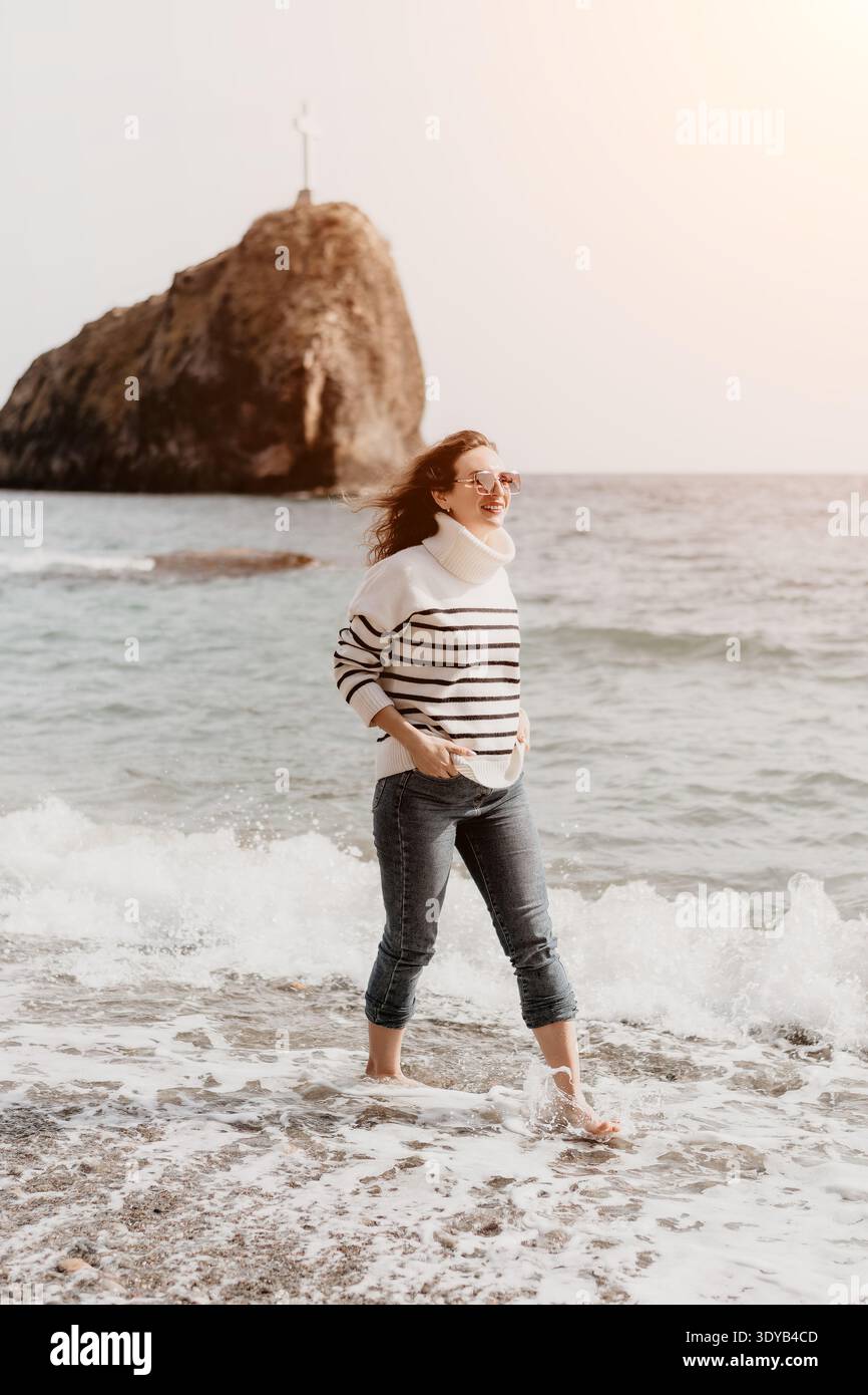 Woman Beach che cammina a piedi nudi attraverso le onde dell'oceano vicino a una formazione rocciosa panoramica con sfondo incrociato durante una vacanza rilassante Foto Stock