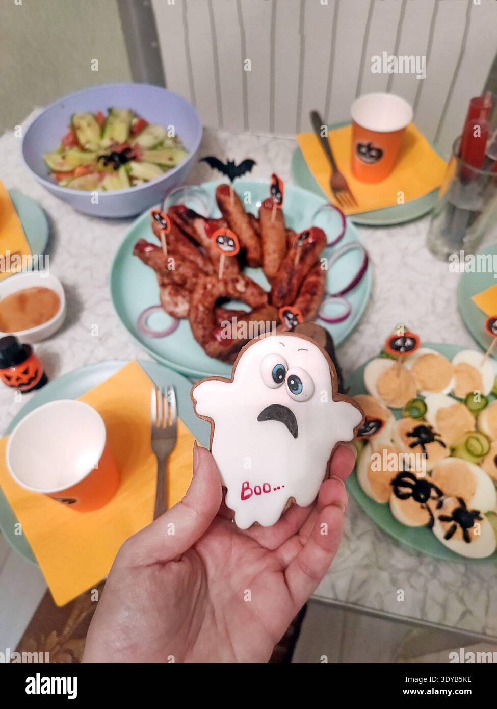 Mano che regge un biscotto di Halloween a forma di fantasma con la parola "Boo", circondato da spuntini festivi, tazze arancioni, salsicce, uova diaboliche e decorazioni Foto Stock