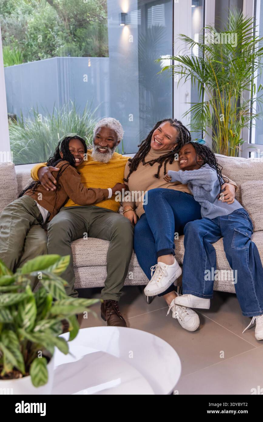 Famiglia afroamericana seduta sul divano beige, ridendo e abbracciata da porte di vetro con maglioni. Casa, calore, insieme, raccolta, luce, interni moderni || modello rilasciato Foto Stock