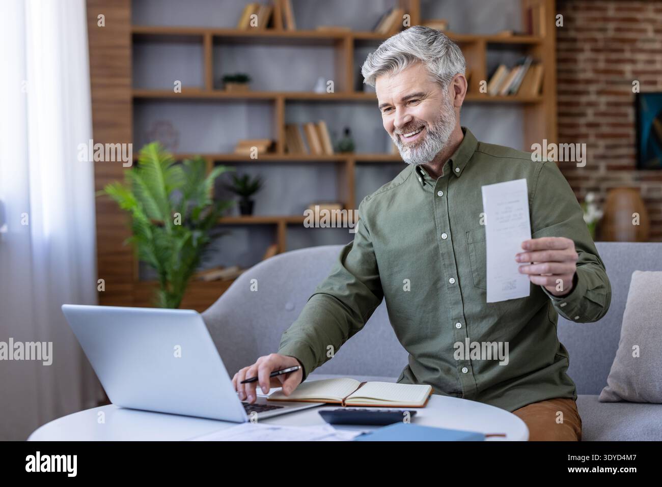 Un dirigente felice che gestisce le finanze domestiche, con una ricevuta cartacea mentre utilizza un notebook e un notebook, dimostrando la pianificazione finanziaria e l'online banking Foto Stock