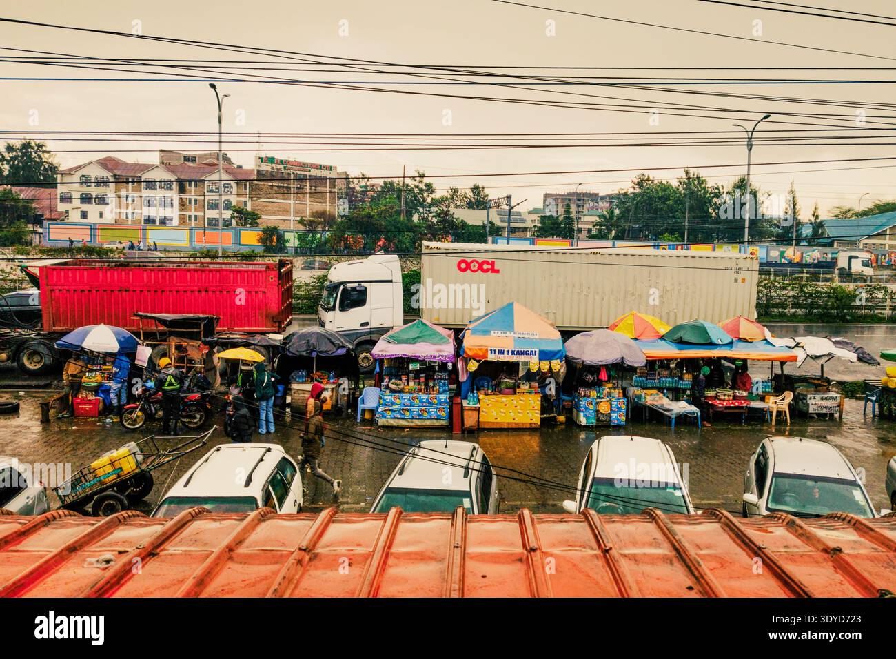 7 marzo 2026 - Rainy Day Roadside Market con camion portacontainer OOCL tra chioschi di veicoli e Boda Bodas a Mlolongo, Kenya, lungo Mombasa Road Foto Stock