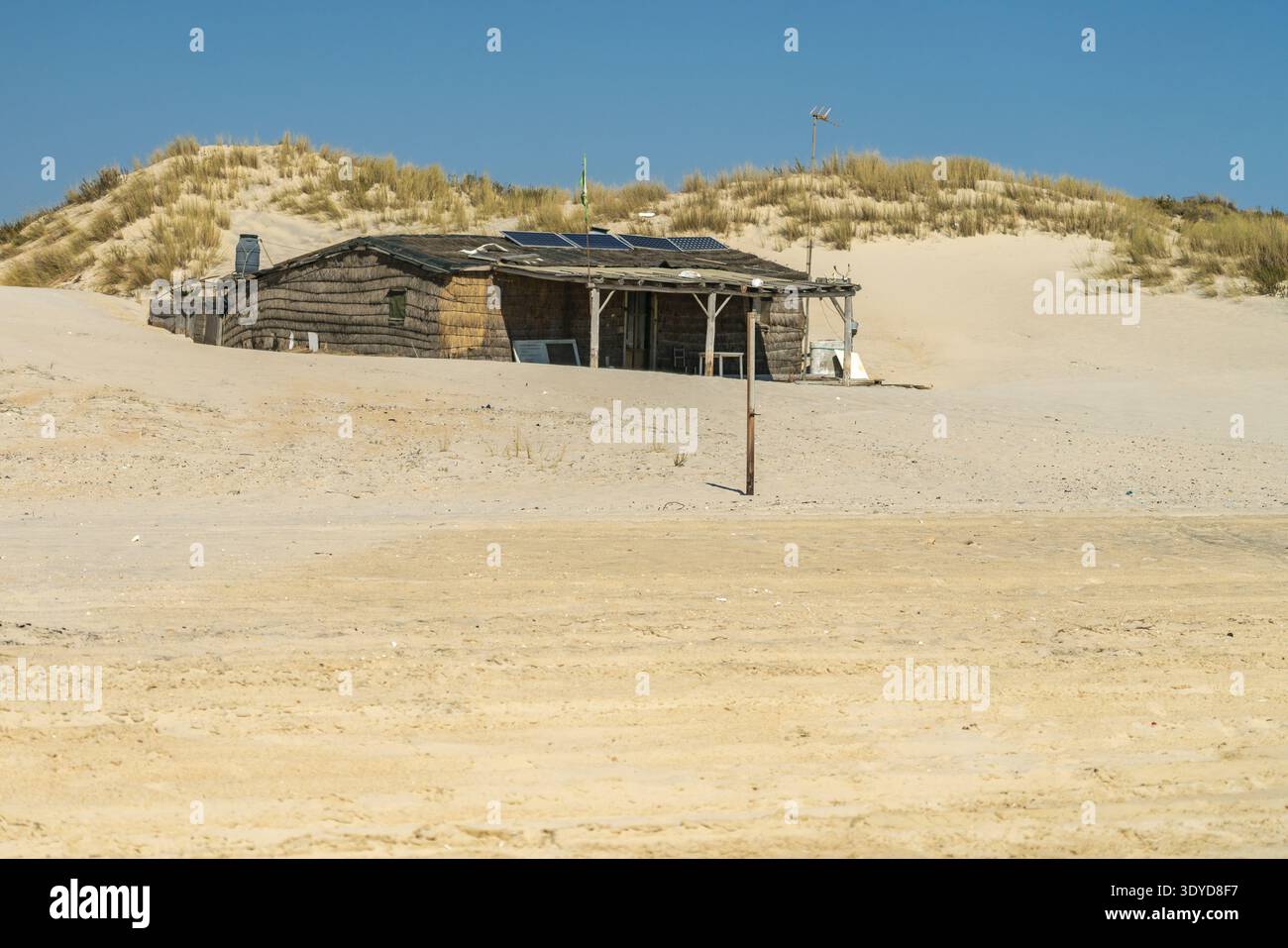 Riparo in legno intempestivo incastonato tra dune di sabbia simili a un deserto, dotato di pannelli solari e di una piccola turbina eolica sotto un cielo azzurro Foto Stock
