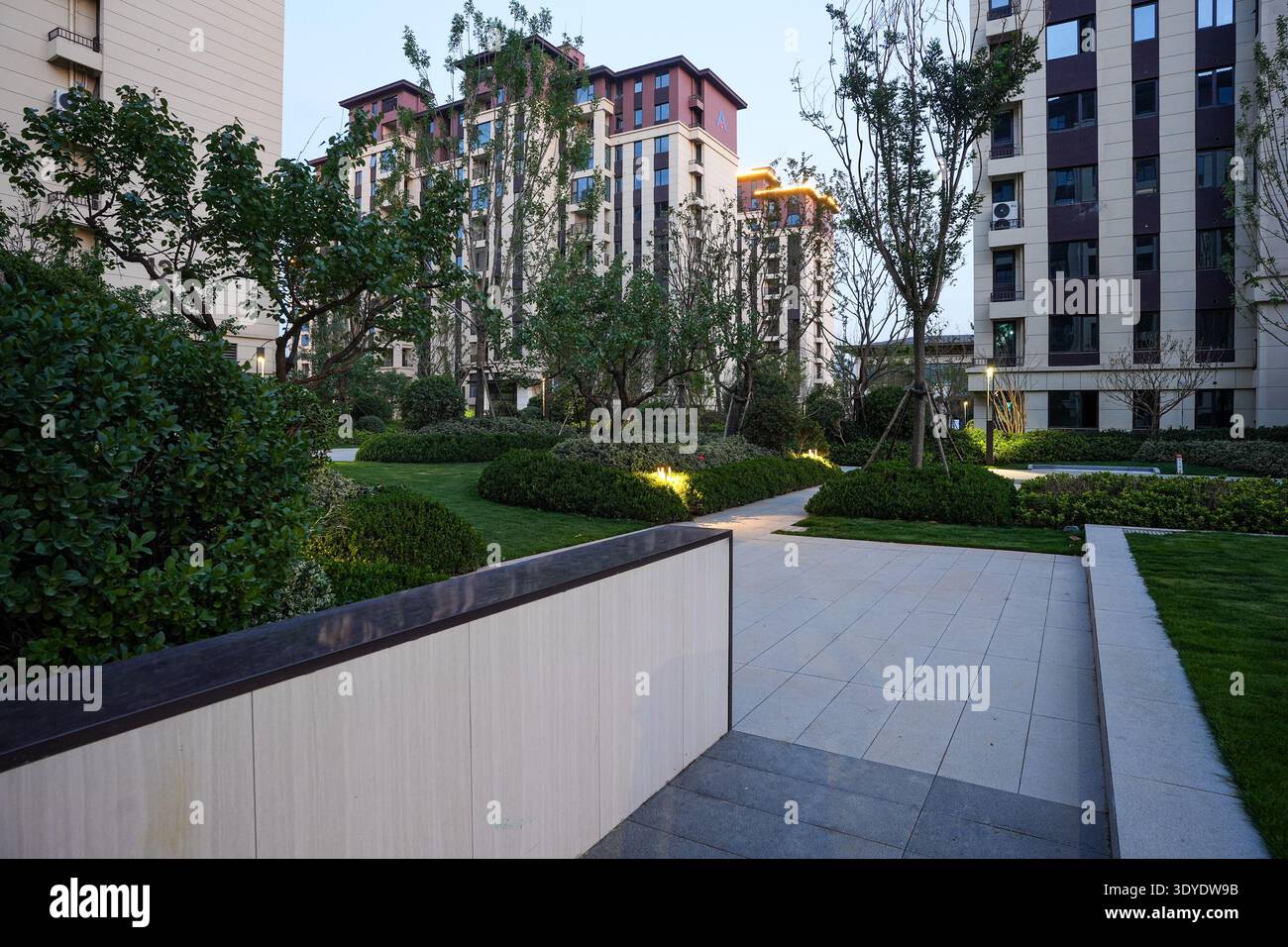Un percorso pedonale strutturato attraverso un parco giardino residenziale al crepuscolo, caratterizzato da luci moderne e lussureggianti arbusti verdi. L'immagine trasmette il leisu serale Foto Stock