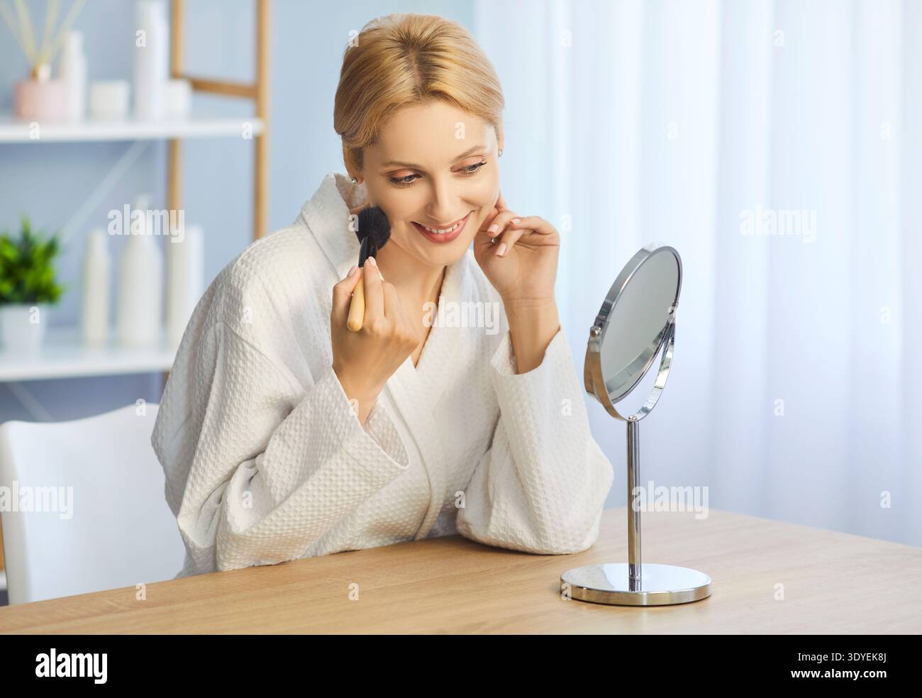 Donna bionda che applica il trucco con un pennello davanti allo specchio Foto Stock
