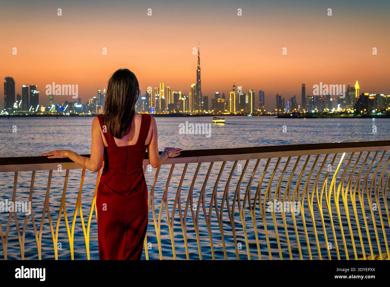 Vista posteriore di una bella donna con un abito da sera rosso che guarda il lontano skyline di Dubai, Emirati Arabi Uniti, durante il tramonto Foto Stock