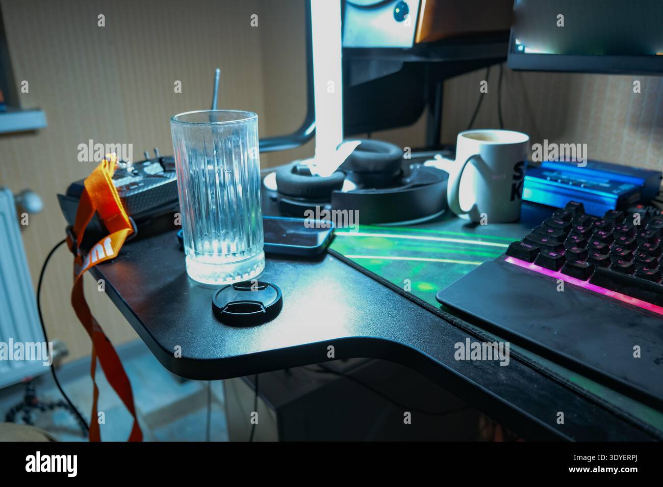 Una scrivania ingombrante in un ufficio domestico bielorusso mostra una tastiera meccanica illuminata in rosa, cuffie su un supporto, una lampada verticale luminosa, una tazza, un telefono, un copriobiettivo, Foto Stock