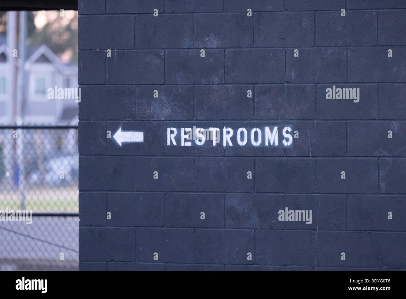 La freccia bianca punta a sinistra sulla parete di mattoni scuri. Il cartello recita "TOILETTE" in grassetto maiuscolo. Lo sfondo mostra un edificio in mattoni dipinto di blu, catena li Foto Stock