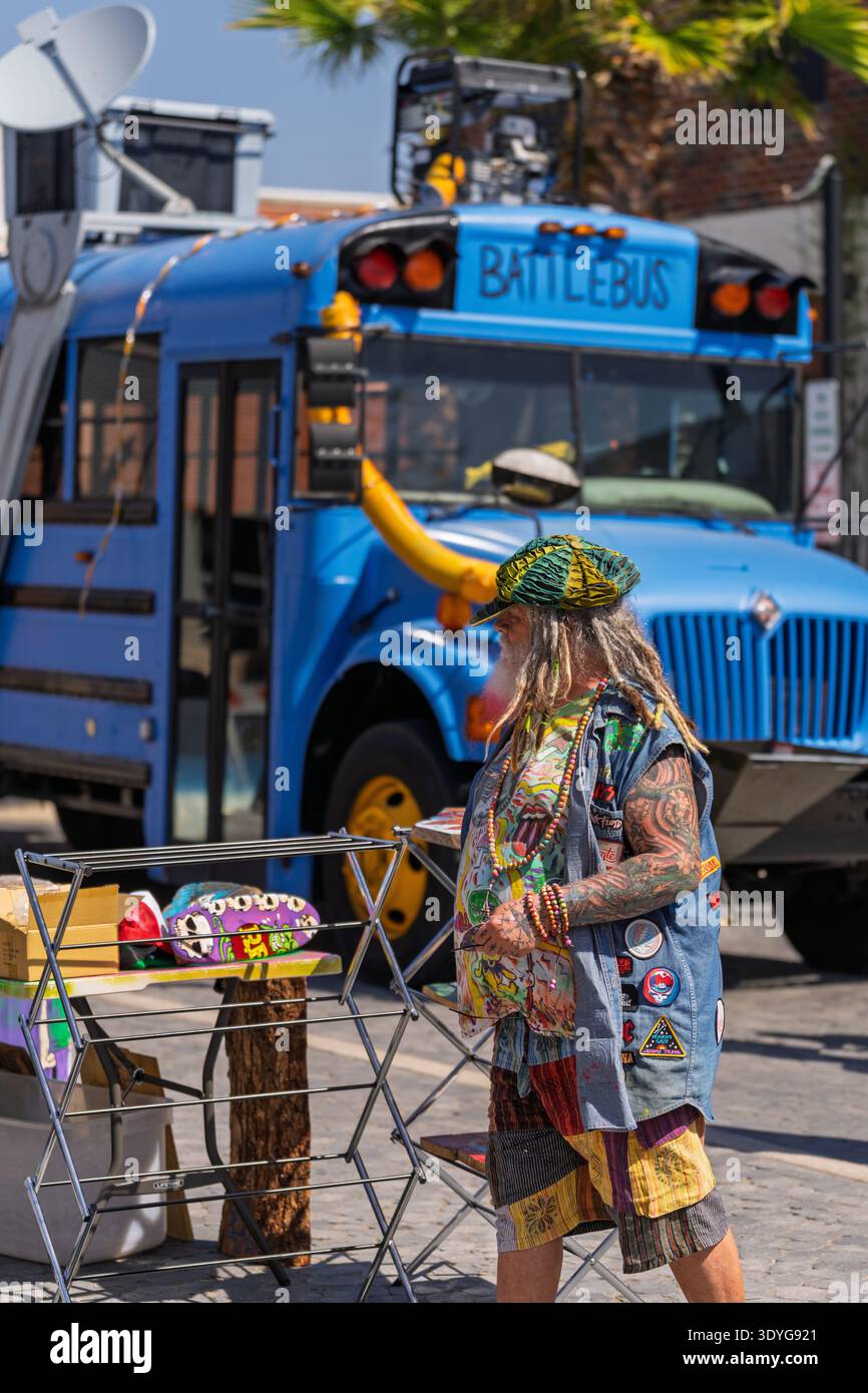 Un uomo con tatuaggi, barba lunga e dreadlocks si trova al suo stand di vendita di fronte a uno scuolabus dai colori vivaci etichettato "Battlebus". Foto Stock