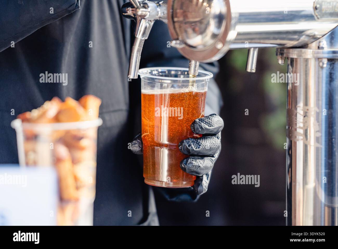 Primo piano di un barista che versa birra alla spina fresca in una tazza di plastica durante un festival all'aperto. Una lager fresca e schiumosa viene servita da una spina Foto Stock