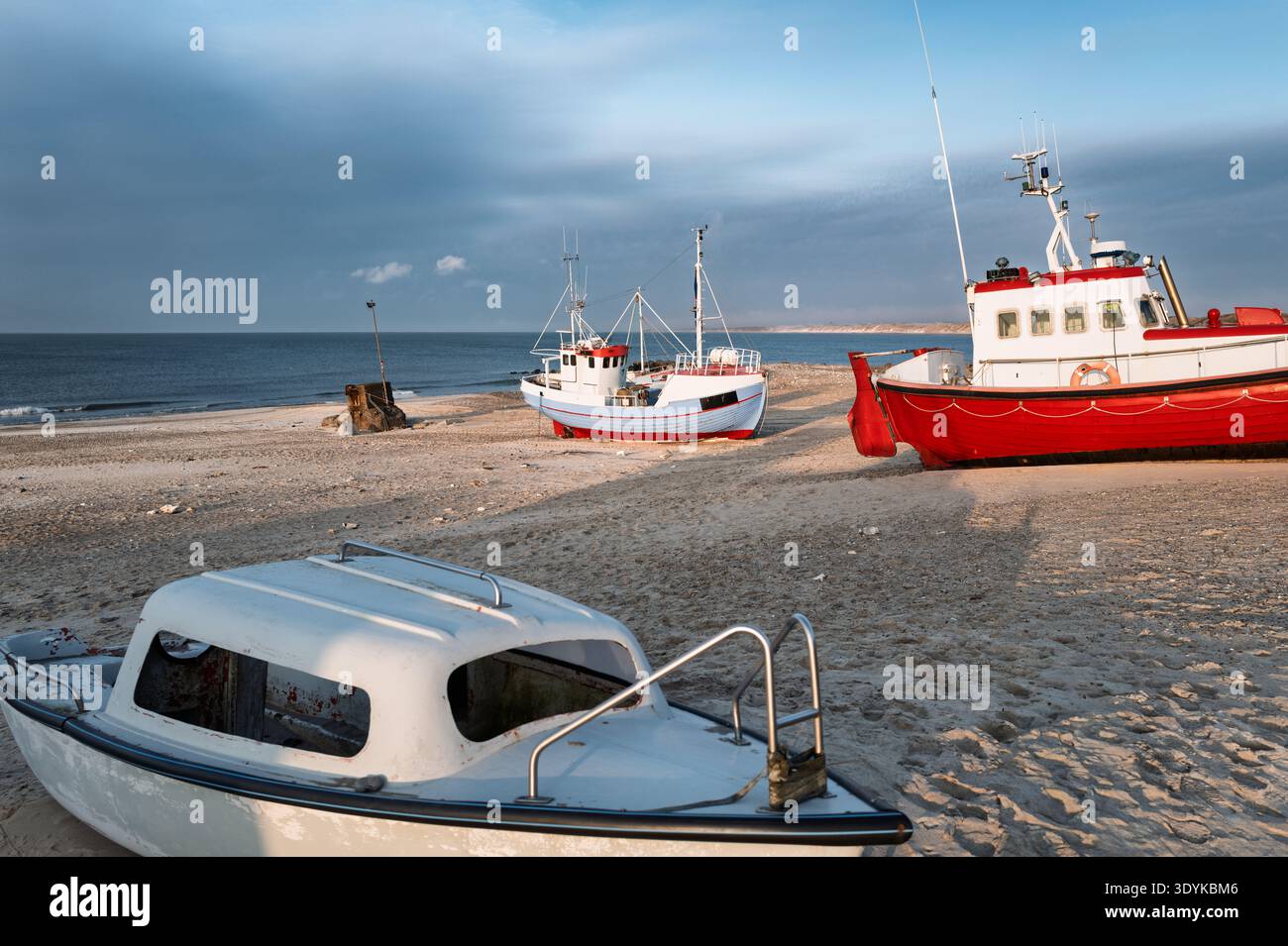 Una colorata barca da pesca siede su di una desolata spiaggia sabbiosa accanto alle dune soffiate dal vento e a una costa calma sotto un cielo grigio coperto, con gabbiani Foto Stock
