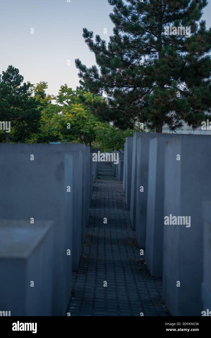Una stretta passerella tra alte stele di cemento a Berlino conduce agli alberi al tramonto. La luce fredda e silenziosa e i verdi tenui fanno da cornice a un punto di svanimento centrale Foto Stock