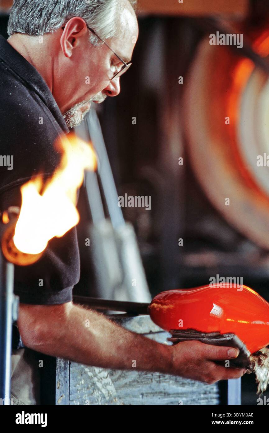 Un vetraio esperto lavora con cura, utilizzando gli strumenti per modellare un pezzo di vetro rosso brillante. L'officina è piena di attrezzature e le fiamme si ergono vicino a. Foto Stock