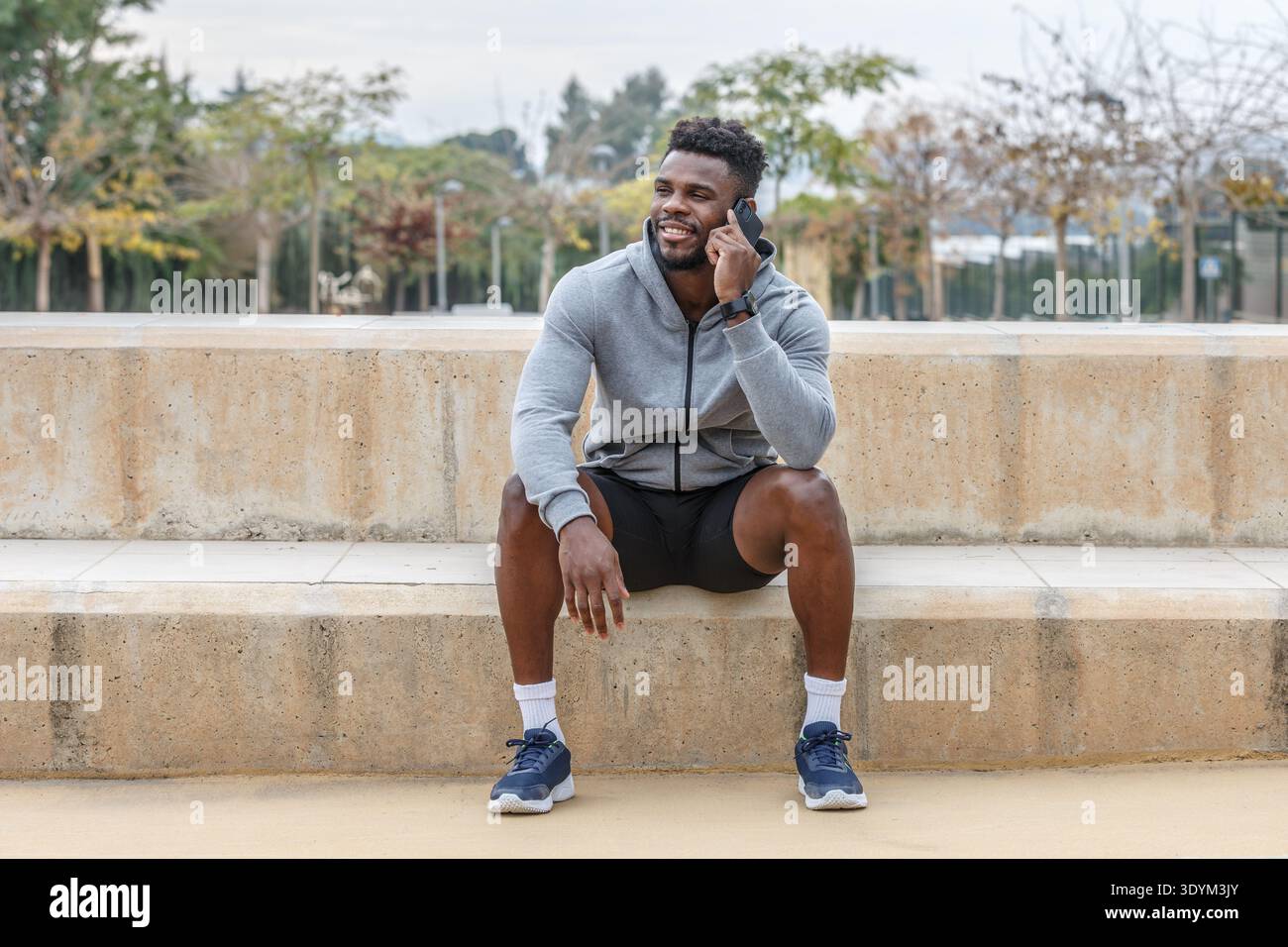 Corpo pieno di giovane atleta afroamericano felice in abbigliamento sportivo seduto sulle scale e a parlare tramite telefono cellulare durante la pausa dall'allenamento Foto Stock