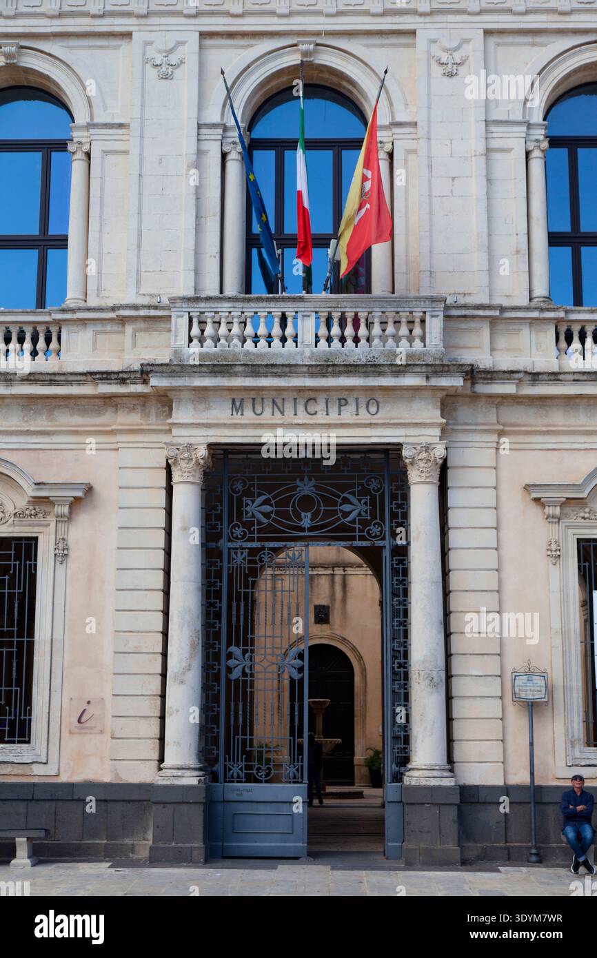Uno storico palazzo municipale italiano dall'aspetto ufficiale con bandiere davanti e un uomo seduto a destra Foto Stock