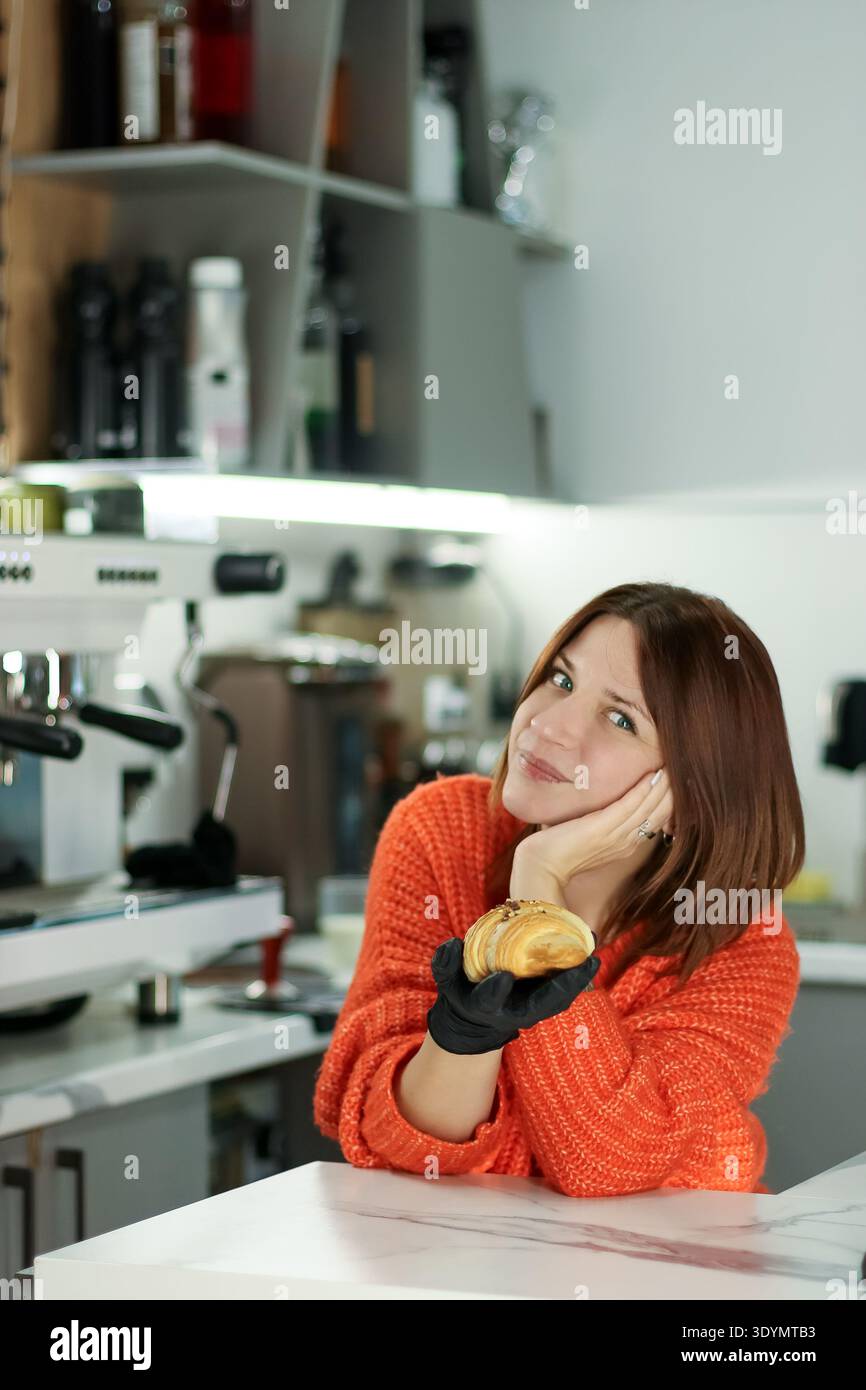 Una donna informale in maglia espone prodotti freschi da forno con guanto igienico all'interno del caffè Foto Stock