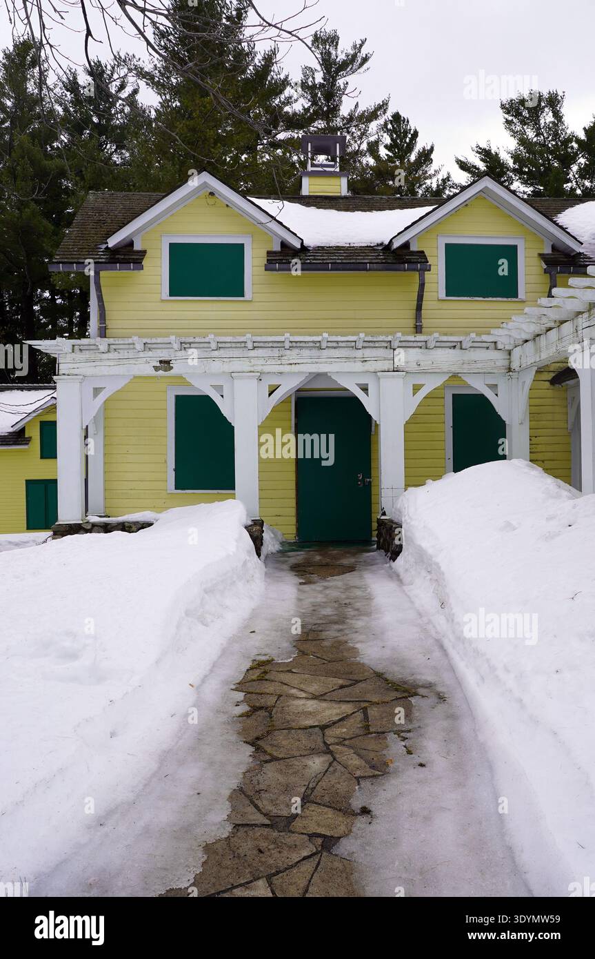Un sentiero in pietra conduce attraverso neve profonda verso l'ingresso di una casa gialla a due piani con persiane verdi e un pergolato bianco in un paesaggio ventoso Foto Stock