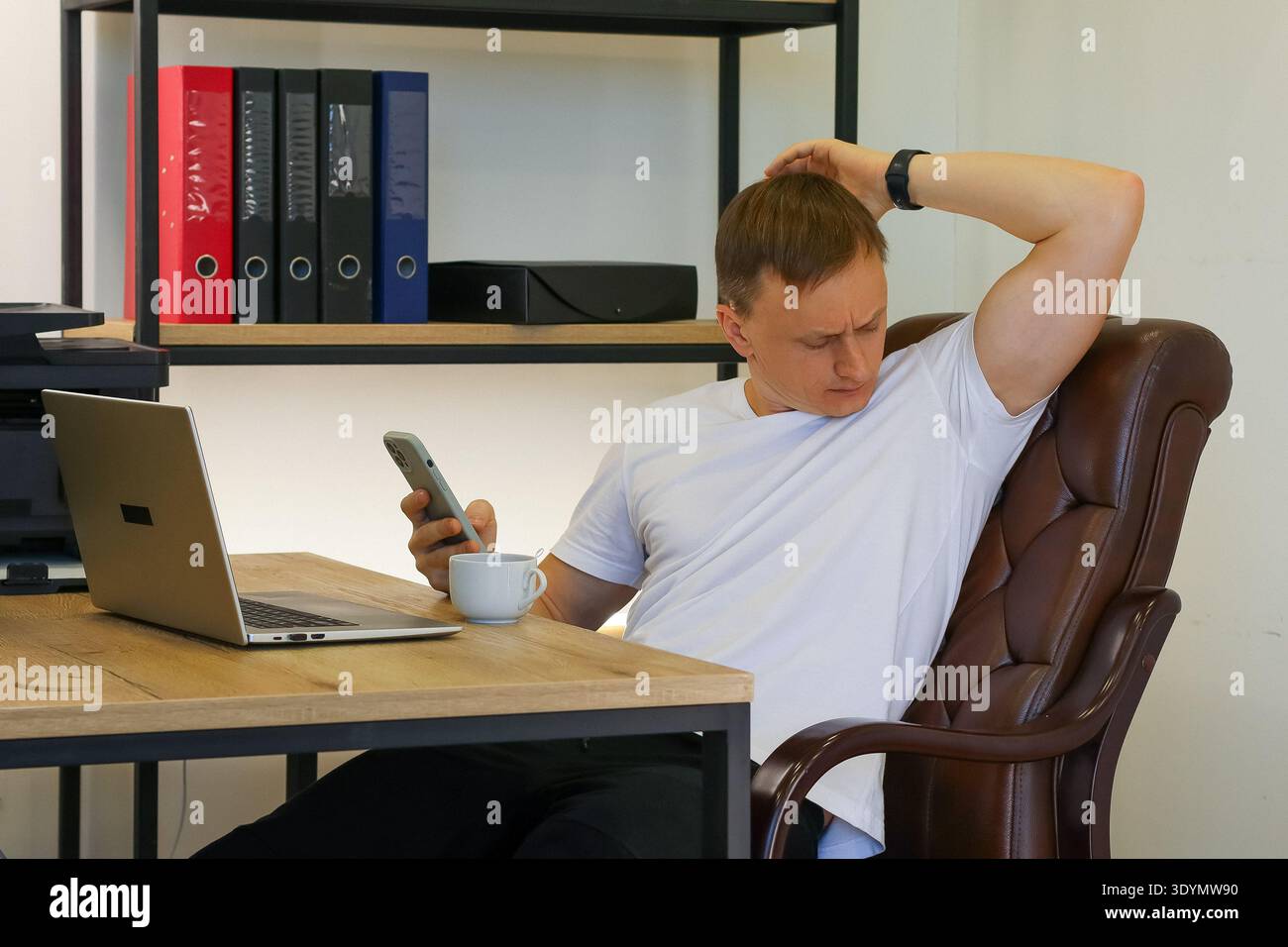 Un lavoratore d'ufficio che si prende cura dello schermo dello smartphone con una tazza di caffè durante le pause sedute nello spazio di lavoro. (92 caratteri) Foto Stock