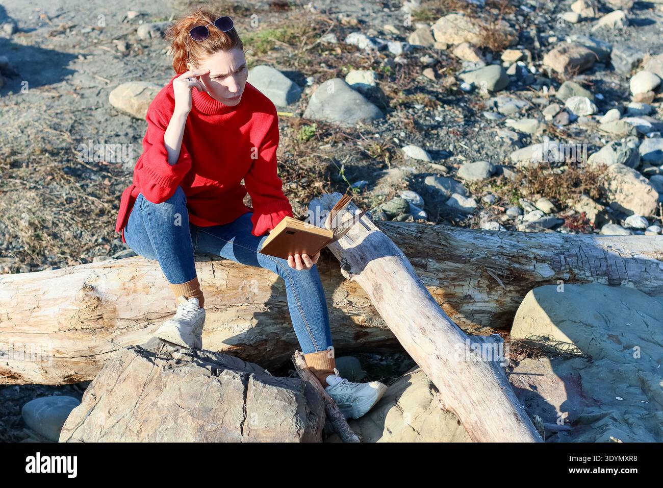 Vista laterale di una giovane donna che tocca il tempio mentre legge un libro su un sedile in legno roccioso all'aperto Foto Stock