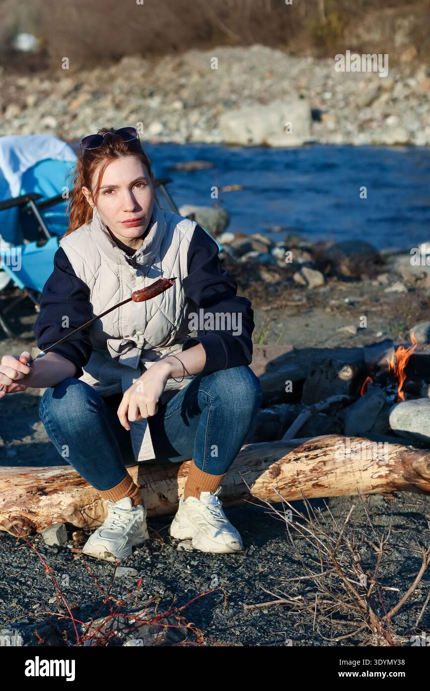 Una giovane donna informale con i capelli rossi tiene la salsiccia spiedata sulle fiamme del fuoco, seduta su tronchi di legno di mare accanto al fiume nel paesaggio della foresta autunnale. Foto Stock