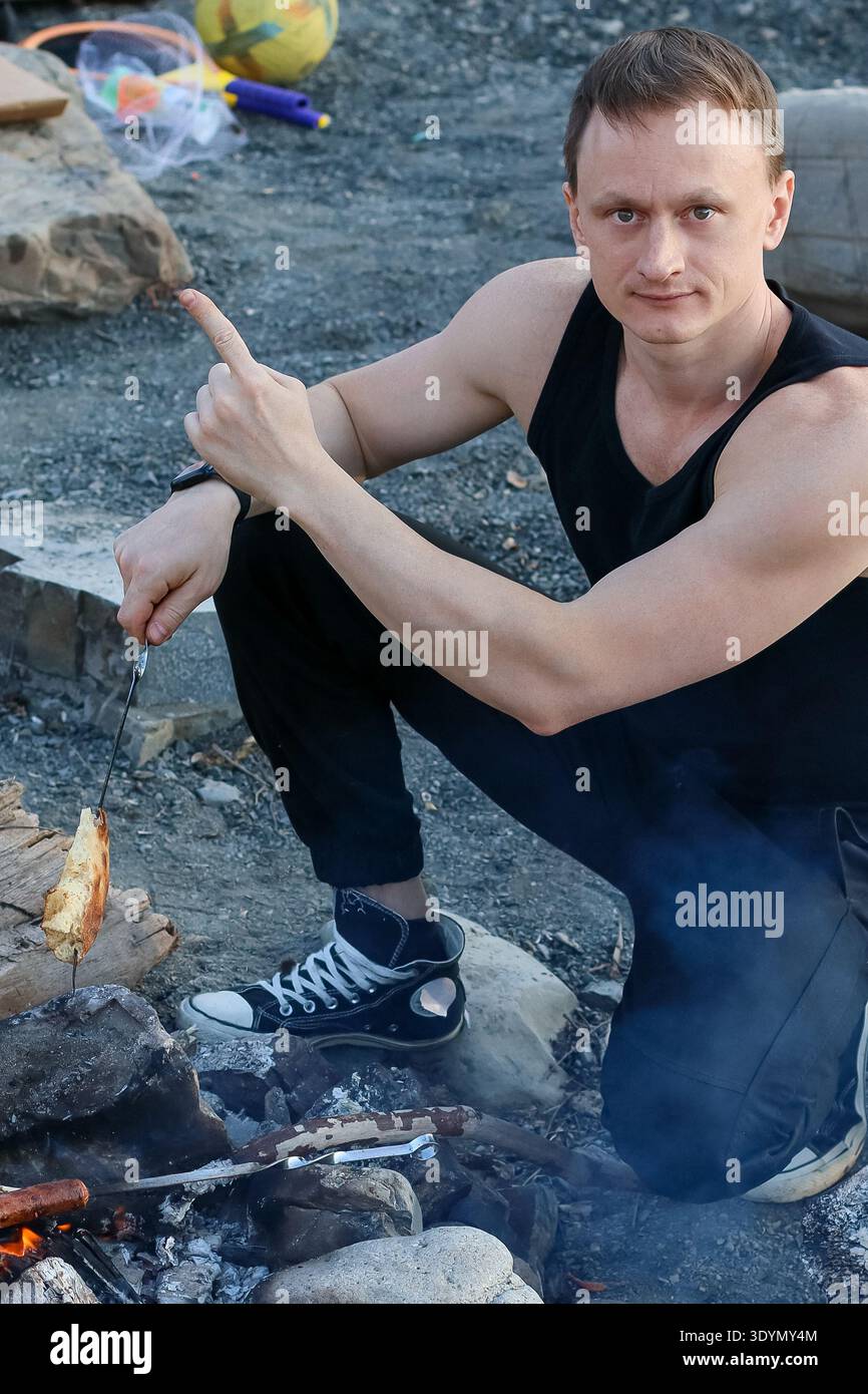 L'uomo in abito nero solleva il dito indice tenendo un pezzo di carne arrosto spietato vicino alle fiamme del fuoco, con cibi da picnic e bevande visibili nell'esterno Foto Stock