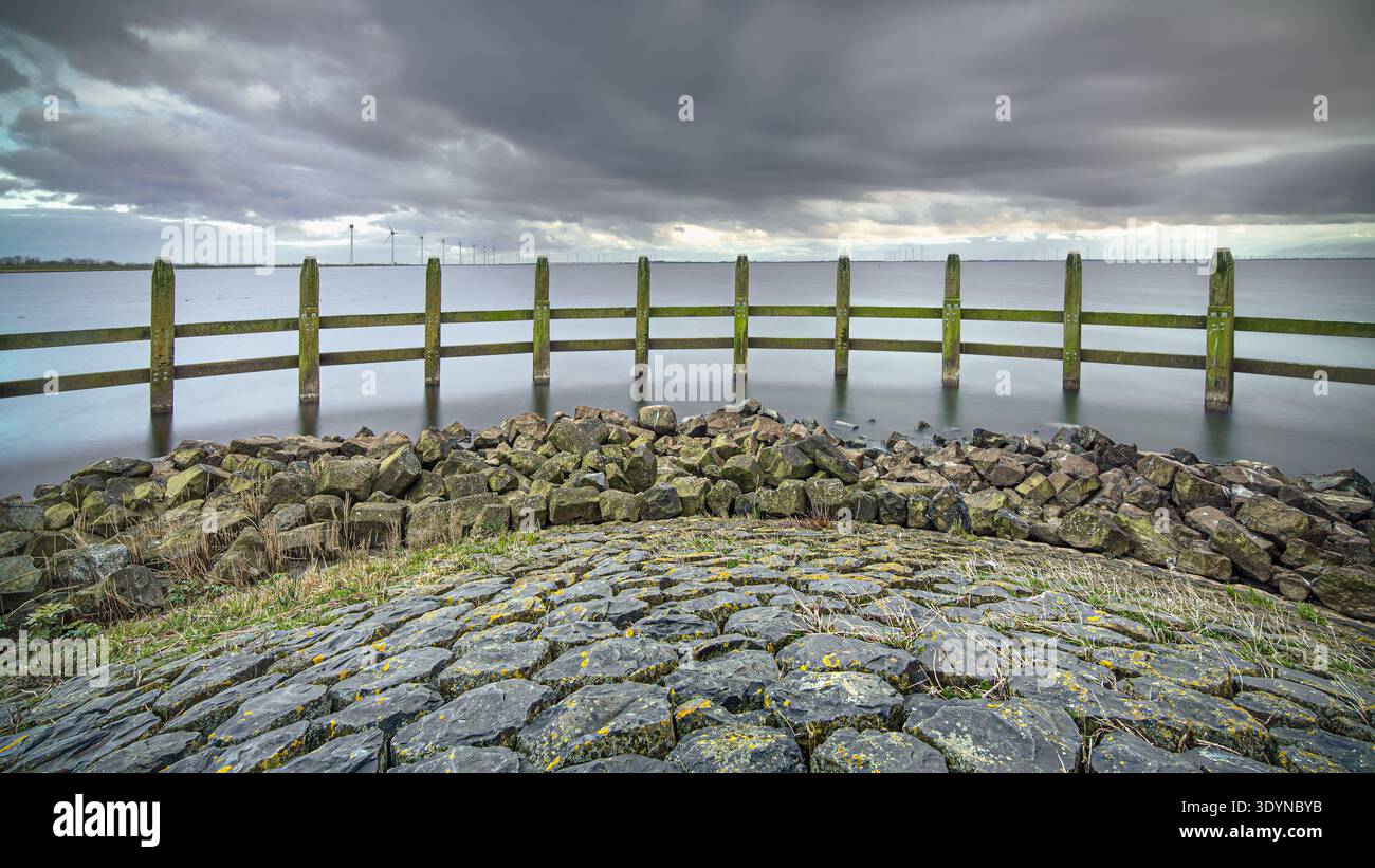 La recinzione in legno si erge a sentinella nelle acque calme vicino a Urk, con turbine eoliche lontane che si stagliano contro un cielo drammatico e nuvoloso. La scena cattura un Foto Stock