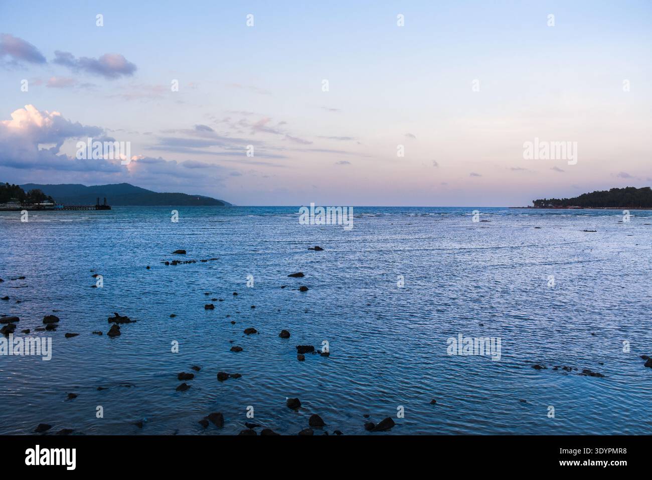 Calmo paesaggio costiero al tramonto caratterizzato da una costa rocciosa, dolci onde e colline lontane sotto un cielo pastello Foto Stock