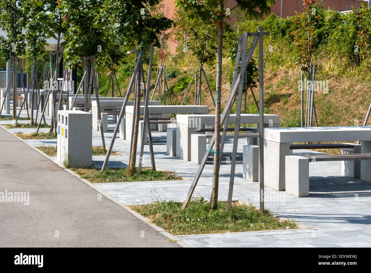 Area di sosta urbana di recente costruzione con tavoli da picnic in granito e giovani alberi lungo la passerella pavimentata nelle giornate di sole. Concetto di sviluppo urbano intelligente e verde Foto Stock
