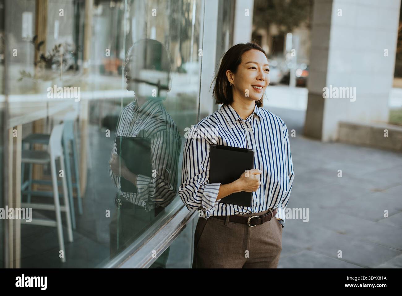 Una donna coreana si trova fuori da un caffè durante il pomeriggio. Tiene una cartella nera e sorride mentre guarda. Il sole si riflette sulla finestra Foto Stock