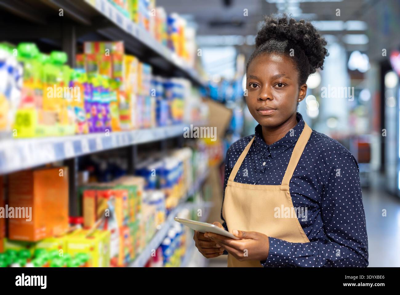 Donna di etnia africana che indossa un grembiule e una camicia a motivi geometrici, in piedi in un corridoio di un negozio di alimentari tra scaffali pieni di vari prodotti, concentrandosi mentre tiene in mano un tablet computer Foto Stock