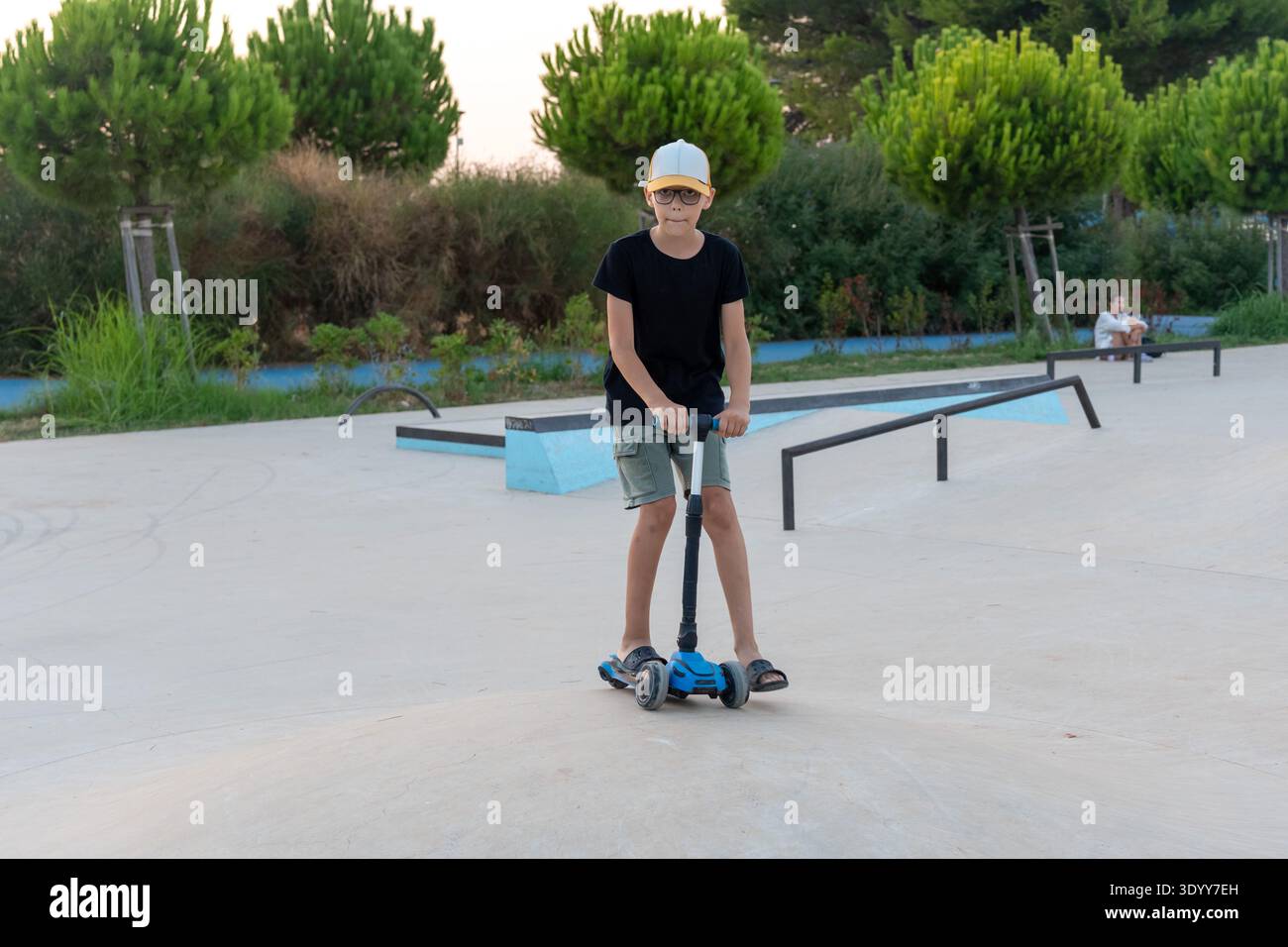 Un bambino sicuro di sé con camicia nera e berretto in posa su uno scooter blu allo skate Park durante l'ora d'oro. Concetto di gioventù urbana fresca, indipendenza e attività Foto Stock