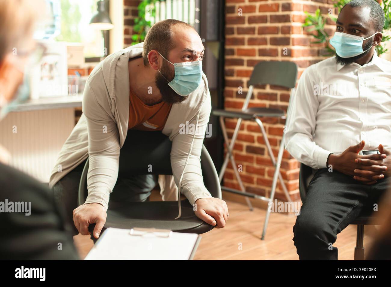 Il giovane in maschera esprime rabbia e disperazione circa la dipendenza da alcol in mezzo alla pandemia. Ragazzo caucasico urlava durante la sessione di gruppo di psicoterapia, avendo un esaurimento nervoso nella stanza delle pareti di mattoni. Foto Stock