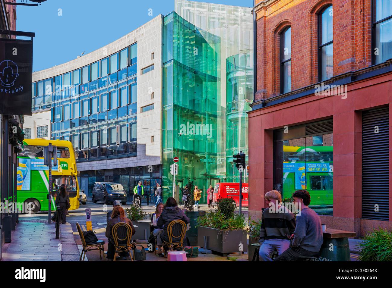 Persone ai tavoli da caffè all'aperto in Stephen Street Lower a Dublino, Irlanda. Moderna architettura in vetro della sede centrale del Dunnes Stores e dell'autobus cittadino. Foto Stock