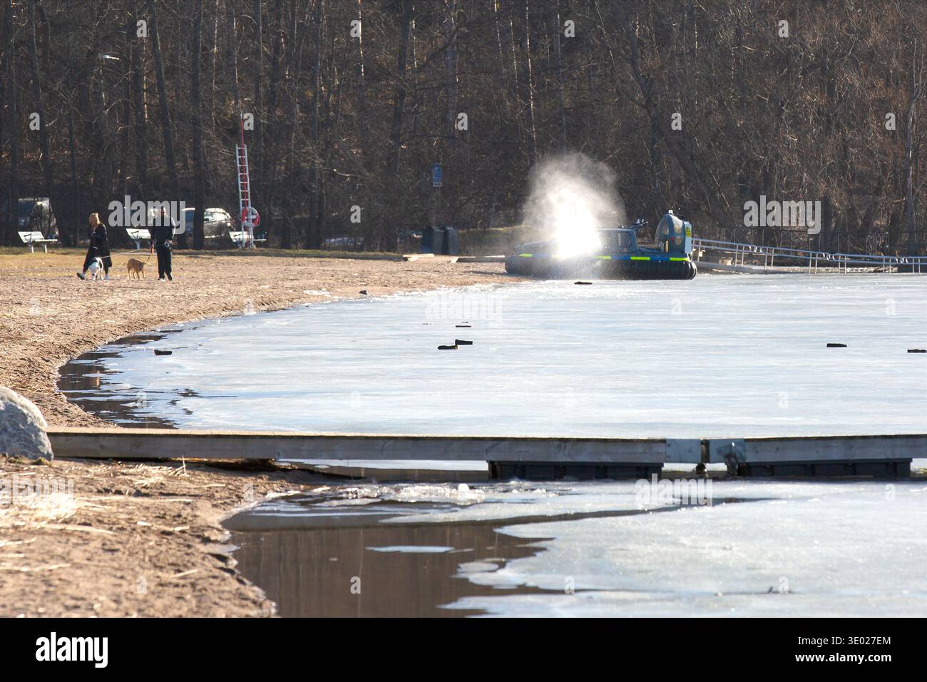 Hovercraft della polizia che opera su un lago parzialmente ghiacciato vicino alla riva mentre le persone camminano con i cani sulla spiaggia di Stoccolma. Foto Stock