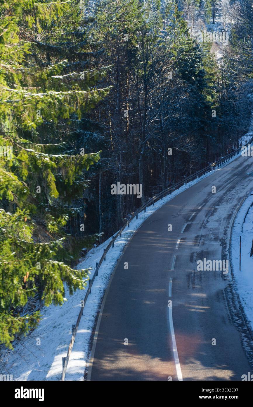 La neve copre il terreno mentre una strada tortuosa serpente attraversa una tranquilla foresta. La luce serale proietta una calda luce sul paesaggio ghiacciato, creando un ambiente sereno Foto Stock
