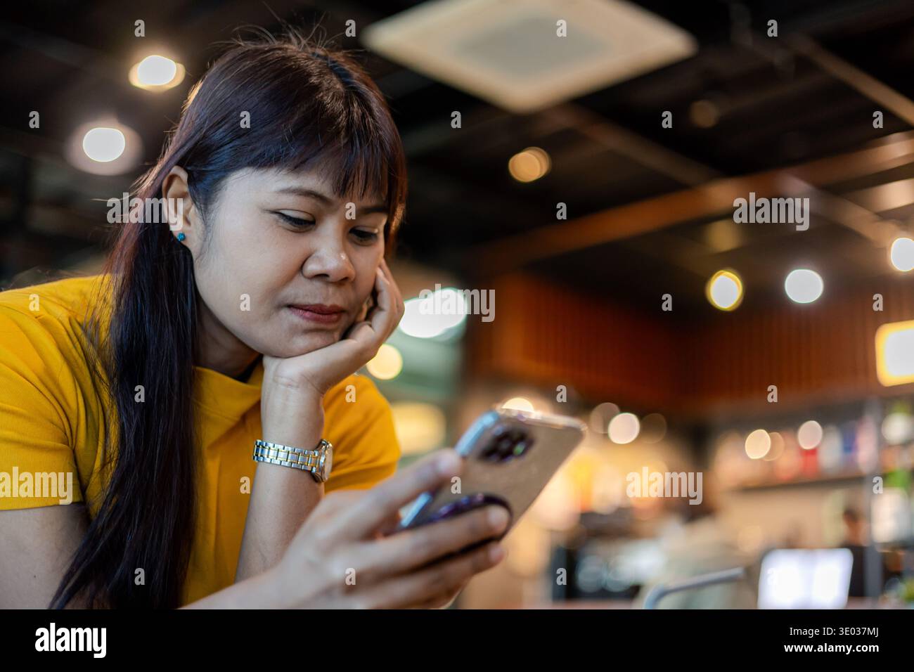 Donna asiatica in maglietta gialla che usa lo smartphone mentre siede in un bar. Concetto di comunicazione mobile, navigazione sui social media, lettura di notizie e digitalizzazione Foto Stock