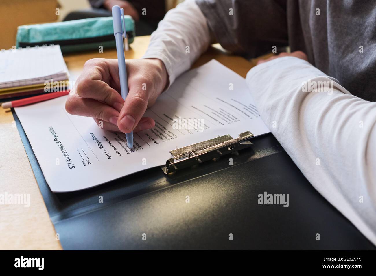 Mano di un adolescente sconosciuto che compila il modulo stampato sugli appunti utilizzando una penna, un quaderno visibile e altri documenti sulla scrivania, indossa una camicia a maniche lunghe, concentrati sull'azione di scrittura Foto Stock