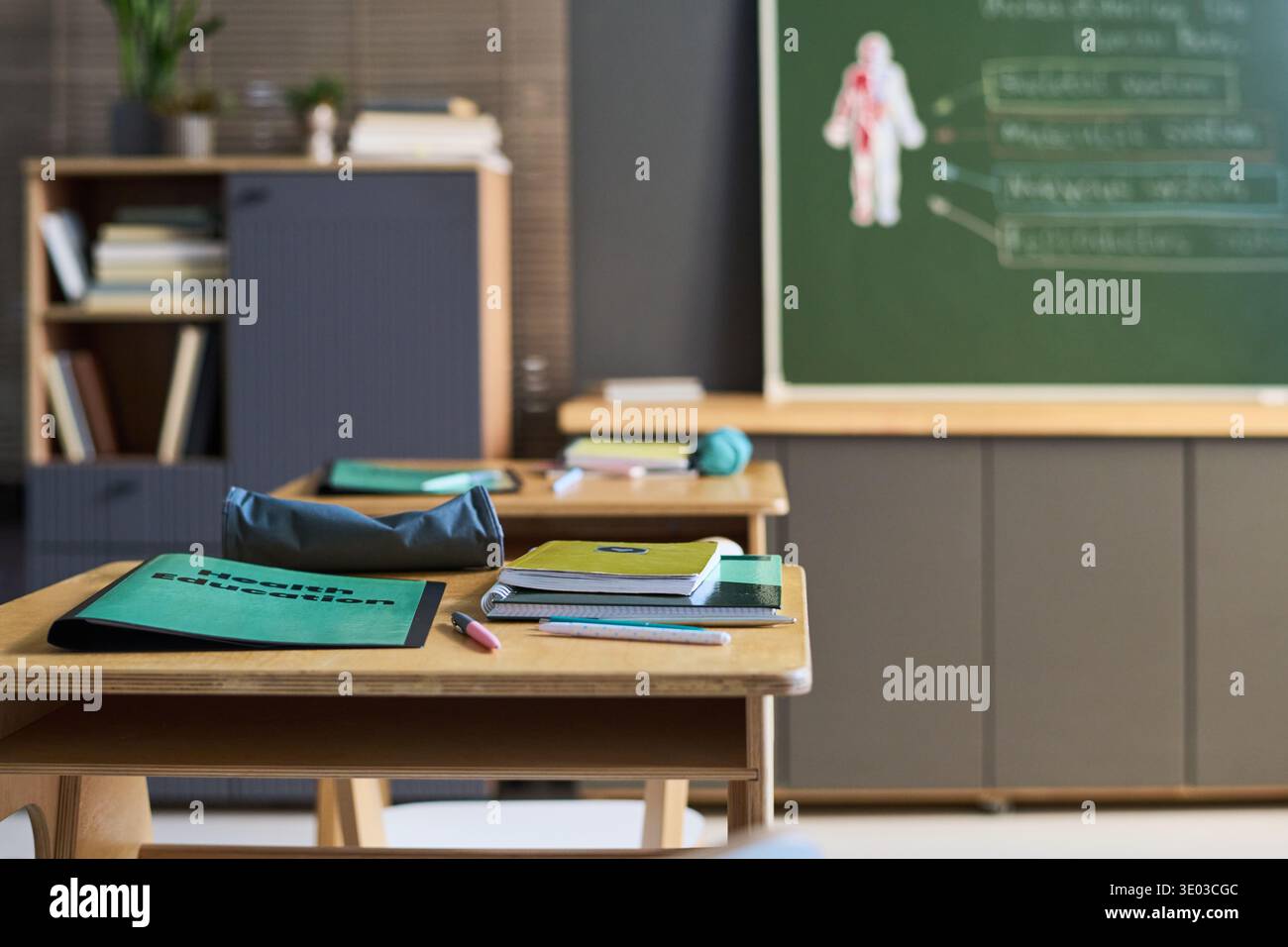 Aula vuota che mostra scrivanie in legno con cartelle di educazione sanitaria, quaderni e matite disposte ordinatamente, lavagna verde con diagramma di anatomia umana visibile sullo sfondo Foto Stock