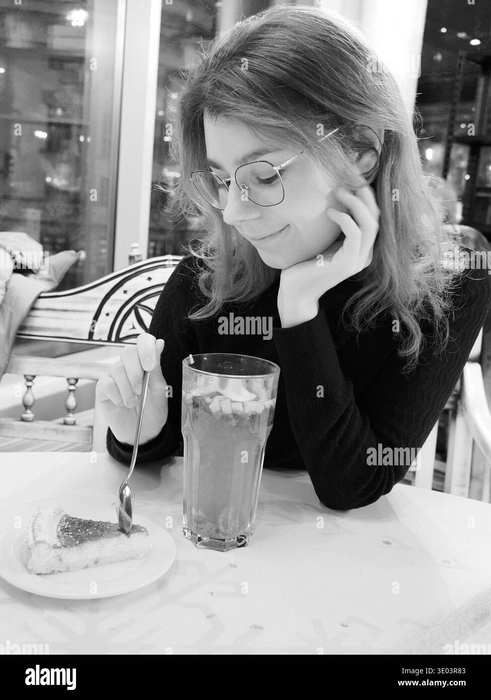 Bianco e nero schietto di una giovane donna in bicchieri che guarda felicemente la sua torta e la sua bevanda calda in un caffè in Estonia Street, Tallinn. Foto Stock